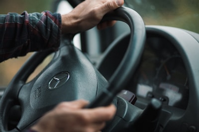 Hands gripping the steering wheel of a vehicle, focusing on the Mercedes-Benz logo prominently displayed at the center. The dashboard and part of the instrument cluster are visible in the background. The lighting appears natural and subdued, hinting at an outdoor setting.