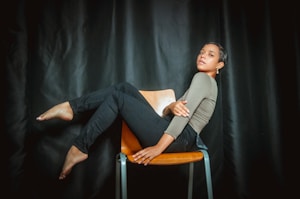 A person is sitting sideways on a wooden chair with their legs extended and crossed, against a dark curtain backdrop. They have a relaxed posture and are looking confidently at the camera, with arms folded across their torso. The lighting casts a soft shadow around them, highlighting the contours of their pose.