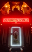 An illuminated shrine with a prominent deity figure surrounded by a bright white light frame. Above the deity is a red neon-lit inscription, with the area cast in a warm, glowing ambience. The setting has intricately designed patterns and pillars, contributing to a sacred and reverent atmosphere.