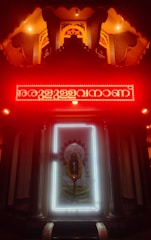 An illuminated shrine with a prominent deity figure surrounded by a bright white light frame. Above the deity is a red neon-lit inscription, with the area cast in a warm, glowing ambience. The setting has intricately designed patterns and pillars, contributing to a sacred and reverent atmosphere.