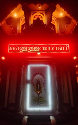 An illuminated shrine with a prominent deity figure surrounded by a bright white light frame. Above the deity is a red neon-lit inscription, with the area cast in a warm, glowing ambience. The setting has intricately designed patterns and pillars, contributing to a sacred and reverent atmosphere.