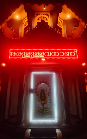 An illuminated shrine with a prominent deity figure surrounded by a bright white light frame. Above the deity is a red neon-lit inscription, with the area cast in a warm, glowing ambience. The setting has intricately designed patterns and pillars, contributing to a sacred and reverent atmosphere.