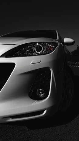 A white car is prominently displayed with a focus on its front headlight and sleek, aerodynamic design. The lighting accentuates the contours and details of the car against a dark background, giving it a dramatic and stylish appearance.