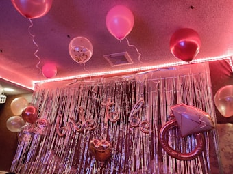 A decorated room features several pink and white balloons with strings, suspended from the ceiling. The background is adorned with metallic silver streamers, creating a shimmering effect. A large inflatable balloon shaped like an engagement ring and a script balloon spell out 'bride to be'. The lighting casts a warm, pinkish glow over the setting.