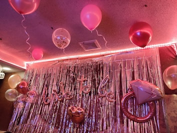 A decorated room features several pink and white balloons with strings, suspended from the ceiling. The background is adorned with metallic silver streamers, creating a shimmering effect. A large inflatable balloon shaped like an engagement ring and a script balloon spell out 'bride to be'. The lighting casts a warm, pinkish glow over the setting.