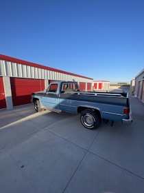 A vintage blue pickup truck is parked in front of red and white storage units under a clear blue sky. The setting appears to be an industrial or storage facility, and the ground is a plain concrete surface.