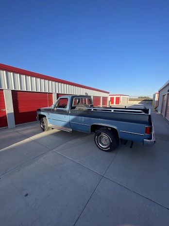 A vintage blue pickup truck is parked in front of red and white storage units under a clear blue sky. The setting appears to be an industrial or storage facility, and the ground is a plain concrete surface.