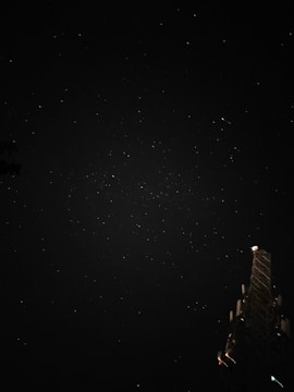 A starry night sky with numerous small white dots scattered across the dark backdrop. In the lower right corner, a large tower-like structure with various antennas and lights is partially visible. The scene suggests a peaceful night environment.