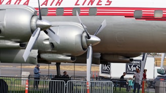 A vintage airplane is displayed with its propeller and engine mechanism visible. Several people are gathered beneath, standing near metal barricades. The plane has a distinctive logo and design, with a classic red and white color scheme. In the background, a catering truck with branding can be seen.