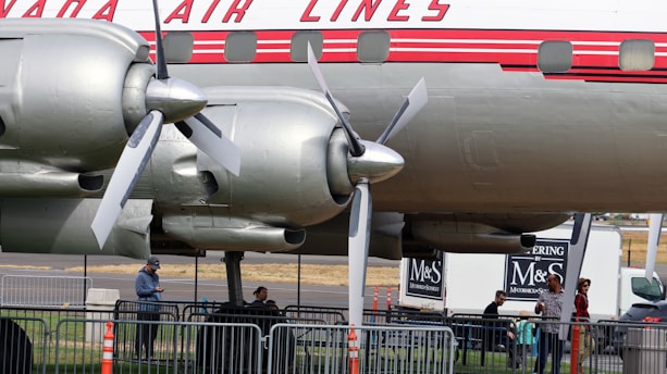 A vintage airplane is displayed with its propeller and engine mechanism visible. Several people are gathered beneath, standing near metal barricades. The plane has a distinctive logo and design, with a classic red and white color scheme. In the background, a catering truck with branding can be seen.