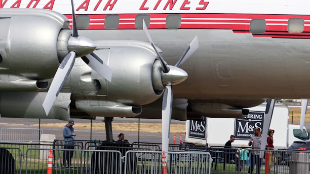 A vintage airplane is displayed with its propeller and engine mechanism visible. Several people are gathered beneath, standing near metal barricades. The plane has a distinctive logo and design, with a classic red and white color scheme. In the background, a catering truck with branding can be seen.