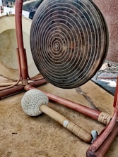 A large metal gong with concentric circles is set up on a wooden frame. In front of the gong lies a mallet with a long wooden handle and a padded head, resting on a brown surface.