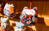 A close-up view of a fabric craft ornament shaped like a cat with a smiling face, adorned with colorful floral patterns. The ornament has a small bell attached to it with a red string and is hanging on a wooden display. Blurred similar ornaments are visible in the background.