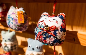 A close-up view of a fabric craft ornament shaped like a cat with a smiling face, adorned with colorful floral patterns. The ornament has a small bell attached to it with a red string and is hanging on a wooden display. Blurred similar ornaments are visible in the background.