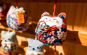 A close-up view of a fabric craft ornament shaped like a cat with a smiling face, adorned with colorful floral patterns. The ornament has a small bell attached to it with a red string and is hanging on a wooden display. Blurred similar ornaments are visible in the background.