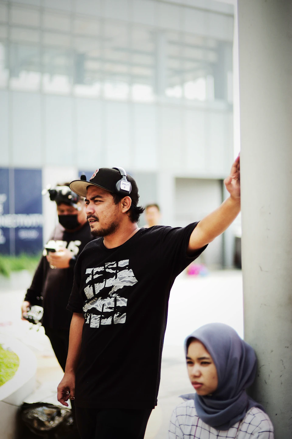 In the image, a man is standing on a sidewalk next to a building with his arm outstretched towards the camera. He's wearing a black t-shirt and a baseball cap. Behind him, there are two other people present - one person in a blue shirt and another person in a white headscarf. The background of the image features a large window that reflects the gray sky above.