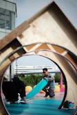 Two individuals are engaged in a creative activity on a vibrant outdoor set with various geometric shapes and colors. One person, wearing sunglasses, is kneeling and handling a piece of teal material, while the other is crouched down holding a device. A structure with a circular opening frames the scene, and a multi-story building is in the background.