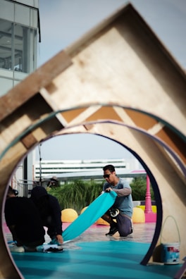 Two individuals are engaged in a creative activity on a vibrant outdoor set with various geometric shapes and colors. One person, wearing sunglasses, is kneeling and handling a piece of teal material, while the other is crouched down holding a device. A structure with a circular opening frames the scene, and a multi-story building is in the background.