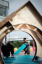 Two individuals are engaged in a creative activity on a vibrant outdoor set with various geometric shapes and colors. One person, wearing sunglasses, is kneeling and handling a piece of teal material, while the other is crouched down holding a device. A structure with a circular opening frames the scene, and a multi-story building is in the background.