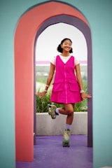 A person is standing on one leg wearing colorful roller skates and a bright pink dress, framed by a multi-colored arched structure. The background features greenery and a soft, overcast sky.