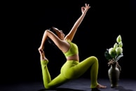 A person in a vibrant green yoga outfit performs a yoga pose, reaching one arm upward and holding a foot with the other. Next to them is a decorative vase with large white flowers and green leaves, set against a stark black background.