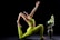A person in a vibrant green yoga outfit performs a yoga pose, reaching one arm upward and holding a foot with the other. Next to them is a decorative vase with large white flowers and green leaves, set against a stark black background.