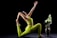 A person in a vibrant green yoga outfit performs a yoga pose, reaching one arm upward and holding a foot with the other. Next to them is a decorative vase with large white flowers and green leaves, set against a stark black background.