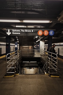 An underground subway station with a sign indicating directions to Uptown, The Bronx, and Queens. The letters B, D, and E are highlighted in colored circles on the sign. Metal railings surround the stairs leading downward, and a few people can be seen in the vicinity.