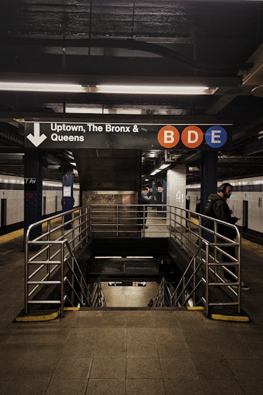 An underground subway station with a sign indicating directions to Uptown, The Bronx, and Queens. The letters B, D, and E are highlighted in colored circles on the sign. Metal railings surround the stairs leading downward, and a few people can be seen in the vicinity.