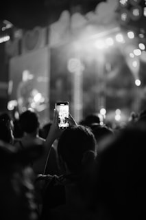 A black and white image featuring a crowd of people at a concert or event. One individual in the foreground holds up a smartphone, capturing the event on camera. Bright lights illuminate the stage in the background, creating a lively atmosphere.