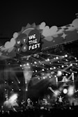 A monochrome image of a music festival stage with bright lights illuminating the scene. Musicians are performing on stage as the crowd watches. The stage design includes a large sign that reads 'We The Fest 2022' with decorative elements.