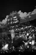 A monochrome image of a music festival stage with bright lights illuminating the scene. Musicians are performing on stage as the crowd watches. The stage design includes a large sign that reads 'We The Fest 2022' with decorative elements.