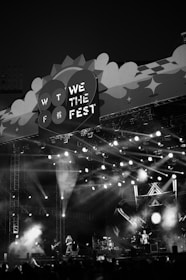 A monochrome image of a music festival stage with bright lights illuminating the scene. Musicians are performing on stage as the crowd watches. The stage design includes a large sign that reads 'We The Fest 2022' with decorative elements.
