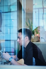 A person is seated indoors, viewed through a transparent glass partition. The individual is holding a smartphone, appearing to be focused and possibly engaged in an activity on the device. There is soft, natural lighting, and elements like a plant and modern decor can be seen in the blurred background.