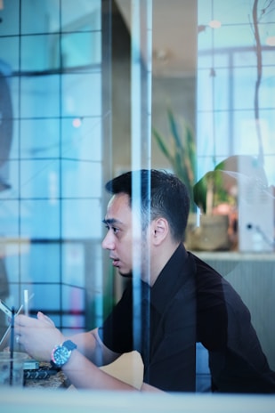 A person is seated indoors, viewed through a transparent glass partition. The individual is holding a smartphone, appearing to be focused and possibly engaged in an activity on the device. There is soft, natural lighting, and elements like a plant and modern decor can be seen in the blurred background.