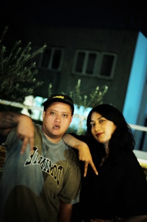 A man and a woman stand together in a dimly lit outdoor setting. The man is wearing a dark cap and a shirt with the word 'DOMINATE' printed on it. He is making a hand gesture. The woman, with long dark hair, is smiling slightly and leaning towards him. There are blurred elements like vegetation and buildings in the background.