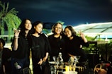 Four women stand close together outdoors at night, smiling and posing for the camera. They are all wearing black outfits and appear to be enjoying each other's company. There are drinks and food on the table in front of them, with a bottle and several cocktail glasses visible. The surrounding environment includes lush greenery and ambient lighting, contributing to a relaxed and joyful atmosphere.