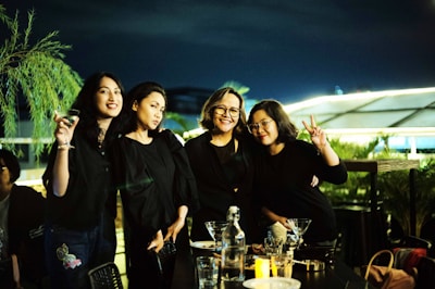 Four women stand close together outdoors at night, smiling and posing for the camera. They are all wearing black outfits and appear to be enjoying each other's company. There are drinks and food on the table in front of them, with a bottle and several cocktail glasses visible. The surrounding environment includes lush greenery and ambient lighting, contributing to a relaxed and joyful atmosphere.
