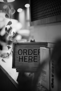 A black and white photo featuring a wooden sign with the words 'ORDER HERE' placed on a countertop next to a plant with hanging leaves. The background is softly blurred, suggesting a cozy café or restaurant ambiance.