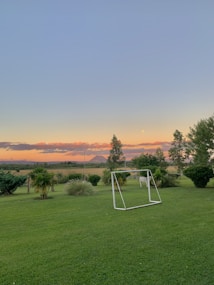 A peaceful landscape featuring a lush green field with a small soccer goal in the center. In the background, there are a few scattered trees and shrubs, with a distant mountain visible under a clear blue sky. The sunset casts a warm orange and pink glow across the horizon, creating a calm and serene atmosphere.