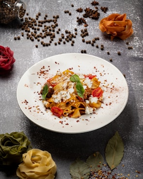 A plate of pasta garnished with cherry tomatoes, fresh basil leaves, and crumbled cheese is placed on a textured gray surface. Surrounding the plate are scattered black peppercorns, star anise, and decorative pasta ribbons artistically arranged to resemble flowers. A few bay leaves and crushed red pepper are also visible, providing a rustic and gourmet presentation.