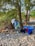 A serene outdoor setting with a backdrop of lush trees and a pebbled ground. A colorful beach towel with a tropical leaf pattern hangs over a branch. Various items are laid out, including a blue inflatable kayak, backpacks, wooden planks, and personal effects such as a towel and water bottle.