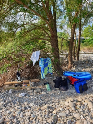 A serene outdoor setting with a backdrop of lush trees and a pebbled ground. A colorful beach towel with a tropical leaf pattern hangs over a branch. Various items are laid out, including a blue inflatable kayak, backpacks, wooden planks, and personal effects such as a towel and water bottle.