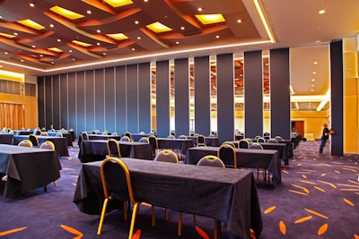 A spacious conference room features neatly arranged tables and chairs covered in black tablecloths. The room has a modern design with strip lighting and a patterned carpet. A person near a foldable wall is adjusting the partitions.