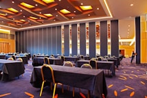 A spacious conference room features neatly arranged tables and chairs covered in black tablecloths. The room has a modern design with strip lighting and a patterned carpet. A person near a foldable wall is adjusting the partitions.
