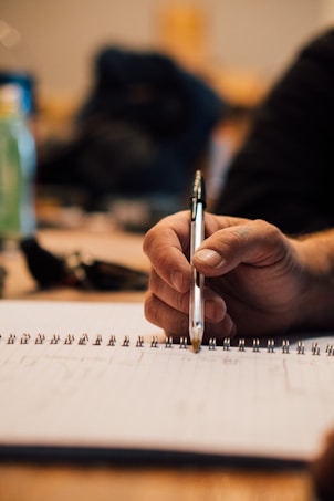 A close-up of a person holding a pen over a spiral-bound notebook, suggesting they are either writing or about to write. The focus is on the hand and the pen, with a blurred background that features indistinct objects.