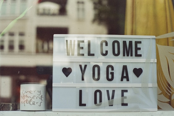 A lightbox sign with the words 'Welcome Yoga Love' is displayed, accompanied by black heart symbols. Beside it, a small white mug bears the handwritten words 'every moment matters.' The background shows partial views of a window with reflections or semi-transparencies of buildings and nature, creating a calming and welcoming atmosphere.