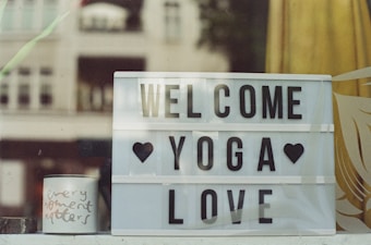 A lightbox sign with the words 'Welcome Yoga Love' is displayed, accompanied by black heart symbols. Beside it, a small white mug bears the handwritten words 'every moment matters.' The background shows partial views of a window with reflections or semi-transparencies of buildings and nature, creating a calming and welcoming atmosphere.