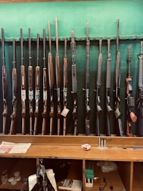 A collection of various rifles is displayed upright against a green felt backing. The rifles have different designs and finishes, featuring wooden and synthetic stocks. Below the rifles, a wooden counter holds miscellaneous items, including a tape measure, some paper, and other small objects.