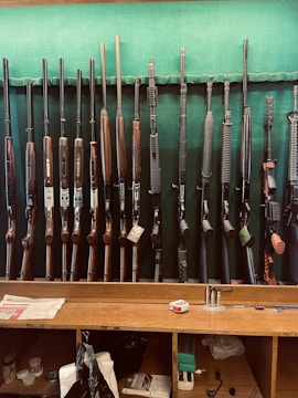 A collection of various rifles is displayed upright against a green felt backing. The rifles have different designs and finishes, featuring wooden and synthetic stocks. Below the rifles, a wooden counter holds miscellaneous items, including a tape measure, some paper, and other small objects.